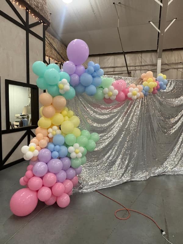 Pastel rainbow balloon garland with white daisy accents framing a silver sequin shimmer wall backdrop, installed by Full of Hot Air in the Kansas City metro