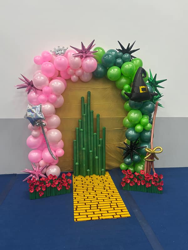 Wizard of Oz themed balloon arch in pink and green with an emerald city and yellow brick road cutout for a school production