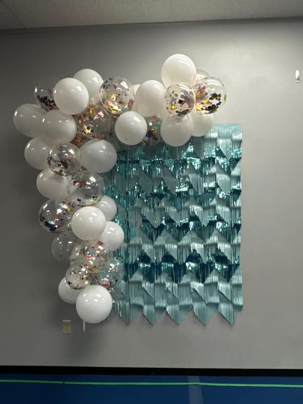 White and confetti balloon garland draped over a teal foil fringe backdrop for a celebration photo wall